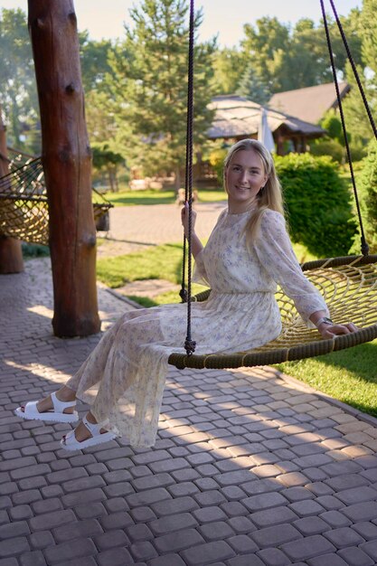 Photo portrait of a young blonde woman in a pastel chiffon dress circling in a wicker hammock