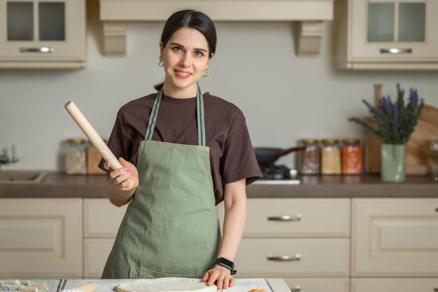 Portrait of a young brunette smiling woman in a kitchen apron with a rolling pin in her hands in a kitchen interior