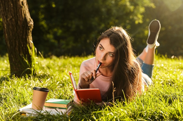 Portrait of young brunette woman thinking and writing down notes in diary while lying at grass in green park on sunny day