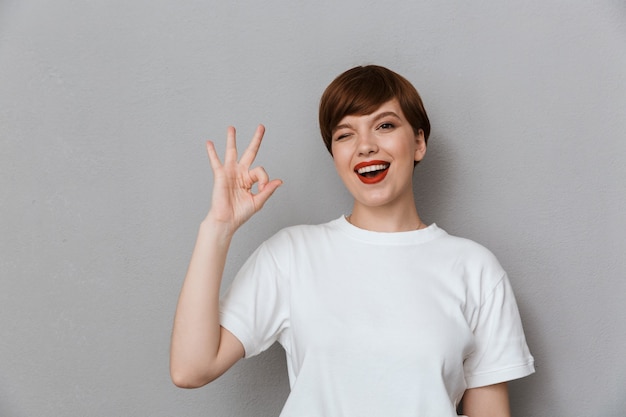 Portrait of young brunette woman wearing casual t-shirt showing ok sign with fingers isolated over gray wall