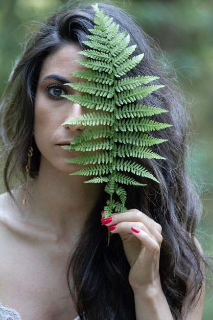 Photo portrait of young brunette woman with a fern leaf covering one eye in the forest