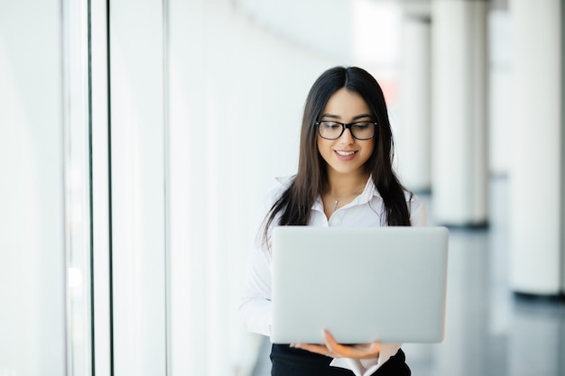 Portrait of young business woman working holding laptop standing against panoramic window with city view