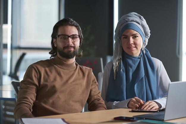 Portrait of young businessman and muslim businesswoman in hijab looking at camera while working at office