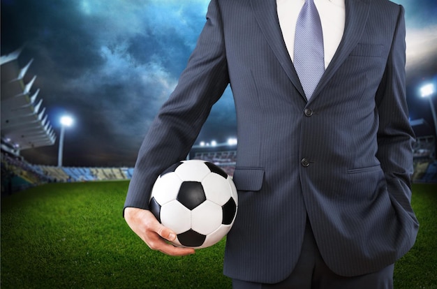 Portrait of young businessman in suit with football ball on stadium background