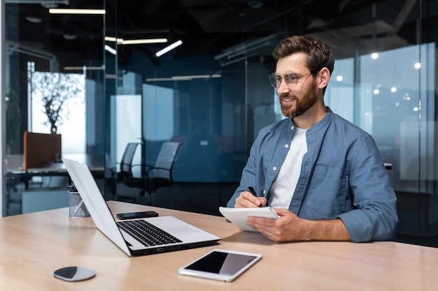 Portrait of a young businessman working in the office using a laptop writing down information in a