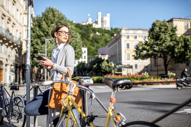 Portrait of a young businesswoman standing with phone near the bicycle on the street in Lyon old town