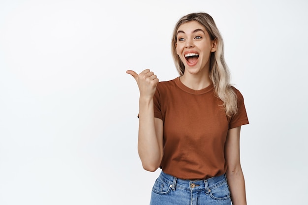 Portrait of young candid woman laughing, pointing and looking left at smth funny, standing in tshirt and jeans on white