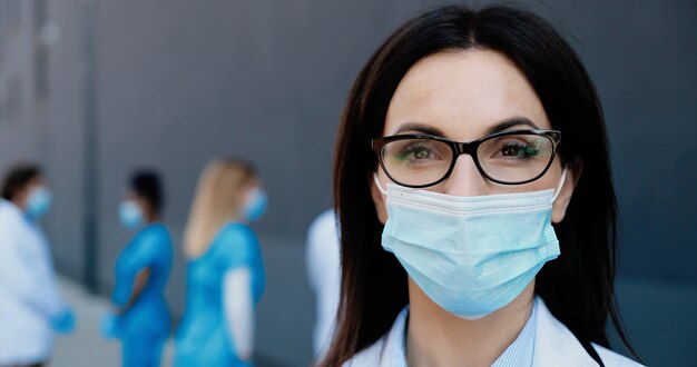 Portrait of young Caucasian brunette beautiful woman doctor in medical mask looking at camera. Close up of female physician in respiratory protection. Mixed-races medics colleagues on background.