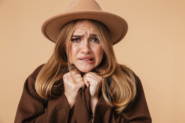 Portrait of young caucasian girl wearing hat and coat trembling and feeling cold isolated on beige