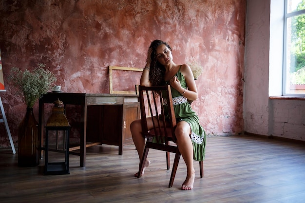 Portrait of a young charming woman with curly hair fairskinned girl in a green dress sits on a chair and with a smile on her face Curly woman in light clothes