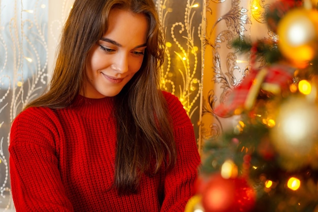 Portrait of a young cheerful girl in bright red sweater decorating Christmas tree