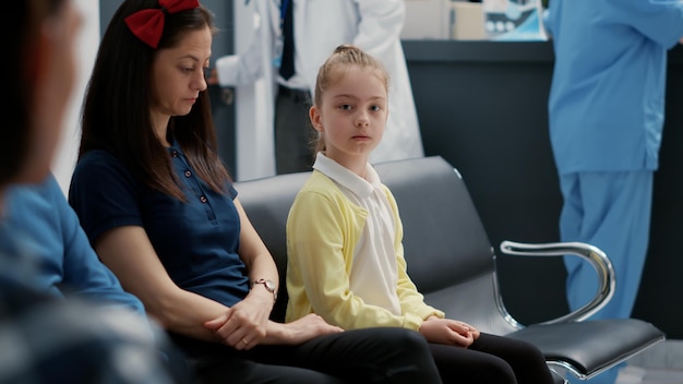 Photo portrait of young child sitting with mother in waiting area at hospital reception, attending annual checkup visit with specialist. little girl in waiting room lobby at healthcare facility.