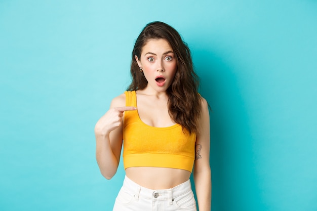 Portrait of young confused woman pointing at herself looking with surprise and disbelief at camera b...