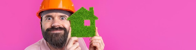 Portrait of young construction engineer wear orange hard hat in a pink jacket standing on red studio background A man holds a green eco house