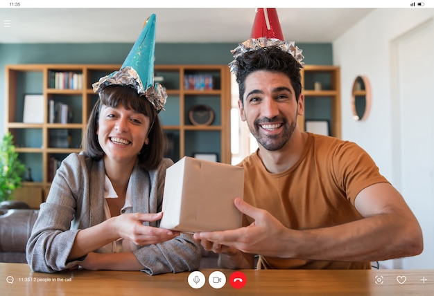 Portrait of young couple celebrating birthday on a video call with gift box from home. New normal lifestyle concept.