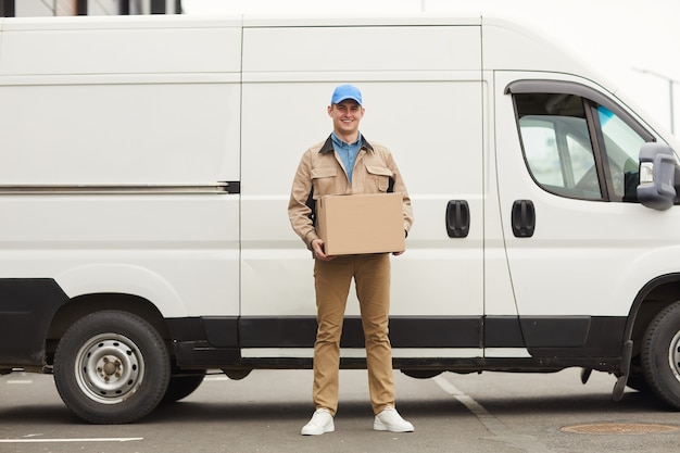 Portrait of young courier holding cardboard box in his hands and looking at camera while standing against the van outdoors