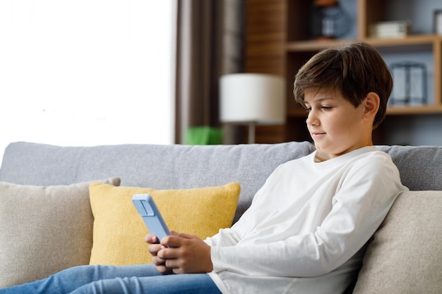 Portrait of young cute boy holding smartphone sitting at sofa Teenager at home choosing favorite music texting messages browsing internet watching video in social media playing online games