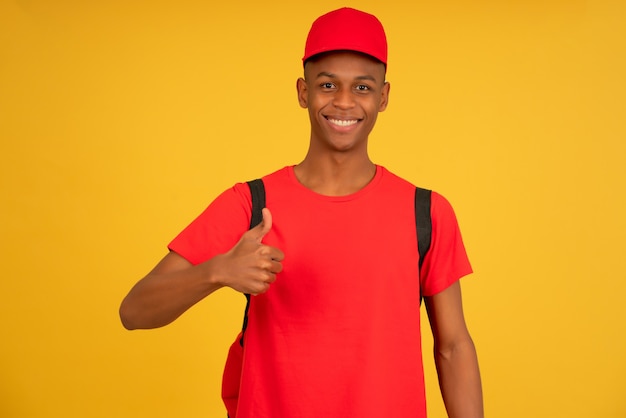 Portrait of a young delivery man looking at the camera and showing thumb up while standing against isolated yellow background. Delivery service concept.