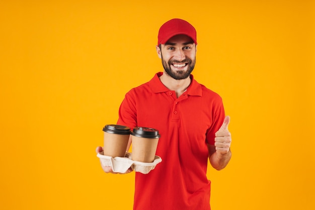 Portrait of young delivery man in red uniform showing thumb up and holding takeaway coffee cups isolated over yellow