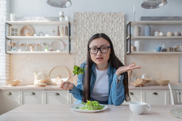 Portrait of young dissatisfied Asian woman eating salad at home and looking at camera