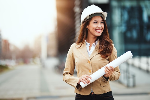 Portrait of young engineer woman with white helmet and blueprints in her hands at construction site.