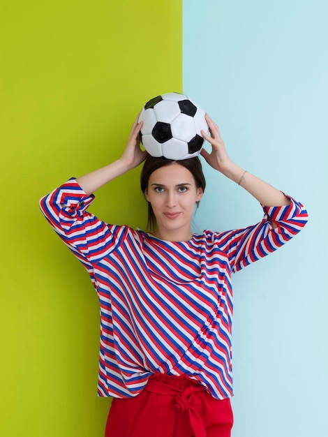 Portrait of young European woman holding soccer ball on her head. Happy girl, football fan or player isolated on green and blue background. Sport, play football, health, healthy lifestyle concept