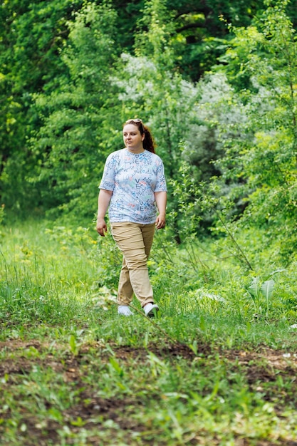 Portrait of young fat woman with long curly red hair walking on path in park forest among green trees Summer weekend