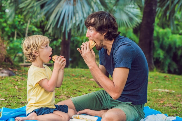 Portrait of a young father and his son enjoying a hamburger in a park and smiling