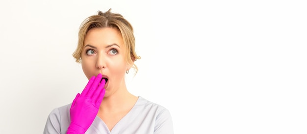 Portrait of a young female caucasian doctor or nurse is shocked covering her mouth with her pink gloved hands against a white background