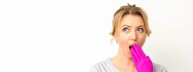 Portrait of a young female caucasian doctor or nurse is shocked covering her mouth with her pink gloved hands against a white background