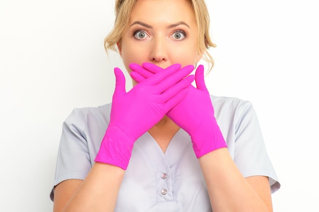 Portrait of a young female caucasian doctor or nurse is shocked covering her mouth with her pink gloved hands against a white background