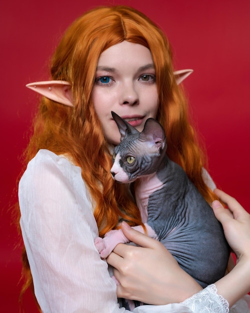 Portrait of young female cosplay elf holding adorable Sphinx kitten in hands on red background