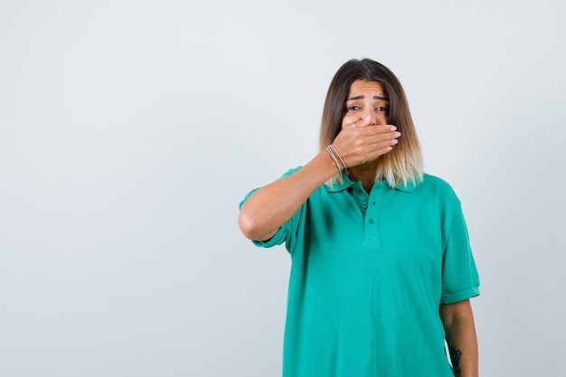 Portrait of young female covering mouth with hand in polo t-shirt and looking shocked front view