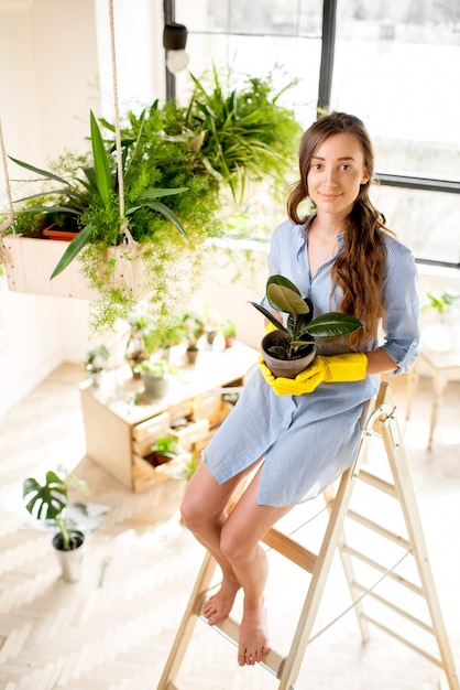 Portrait of a young female gardener holding a flowerpot sitting on the ladder in the orangery