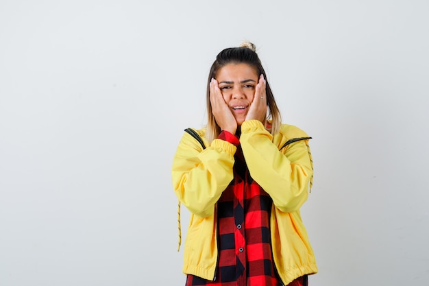 Portrait of young female leaning cheeks on palms in checkered shirt, jacket and looking glad front view