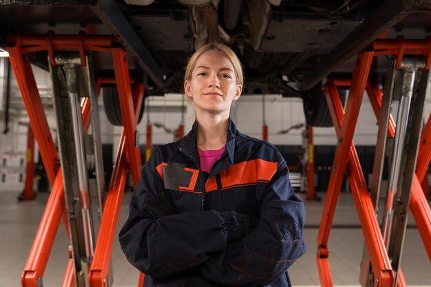 Portrait of a young female mechanic in uniform who looks into the camera against the background of cars being repaired at a service station