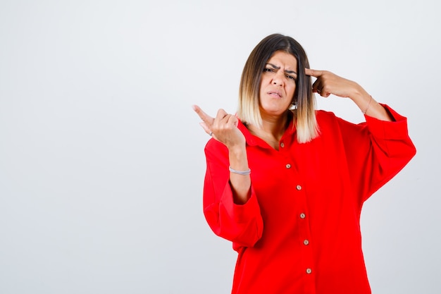 Portrait of young female pointing at head and aside in red oversized shirt and looking hesitant front view