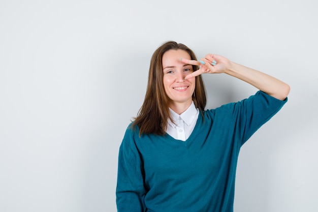 Portrait of young female showing V-sign on eye in sweater over shirt and looking cheery front view