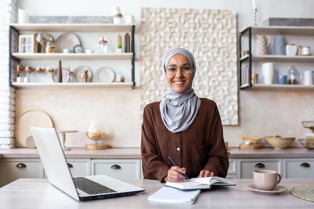 Portrait of a young female student of oriental origin in a hijab sitting at home in the kitchen and