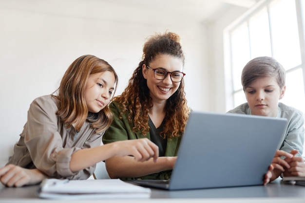 Portrait of young female teacher using computer with children