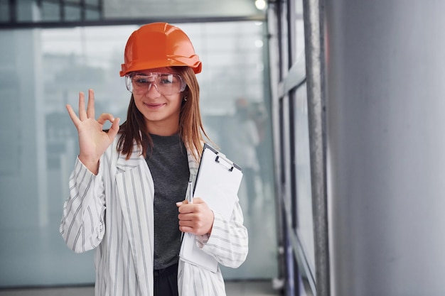 Portrait of young female worker with protective helmet, eyewear and notepad. Standing in the office.