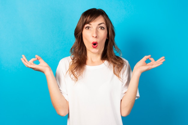 Portrait of a young friendly woman in casual t-shirt with a gesture of meditation and a surprised face on blue, relax