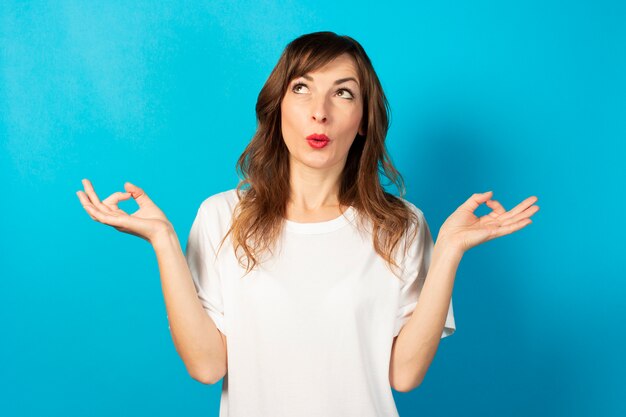 Photo portrait of a young friendly woman in casual t-shirt with a gesture of meditation and a surprised face on blue, relax