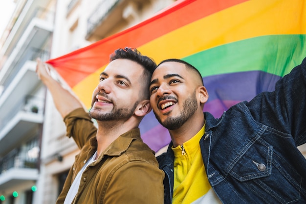 Portrait of young gay couple embracing and showing their love with rainbow flag at the street. LGBT and love concept.
