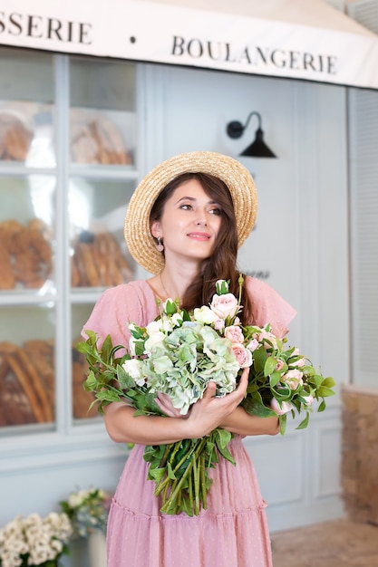 portrait young girl in dress and straw hat holding bouquet of flowers against french bakery