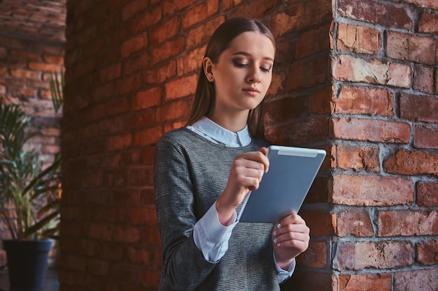 Portrait of a young girl dressed in an elegant gray dress leaning against brick wall, using the tablet.