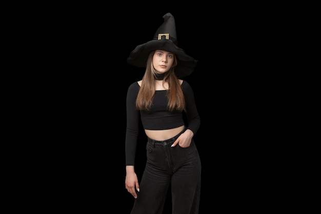 Portrait of young girl in pointed witch hat and black clothes is isolated on black background Copy space Halloween costume