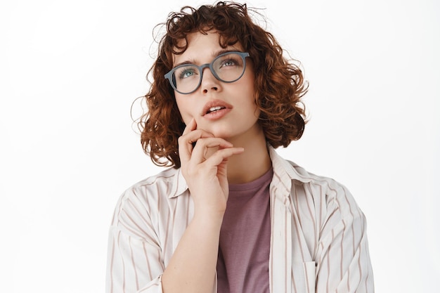 Portrait of young girl student thinking, woman in glasses looking up thoughtful, pondering, solving puzzled, staring at top advertisement and makign choice, white background