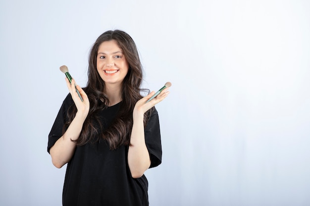 Portrait of young girl with cosmetic brushes looking at camera on white.