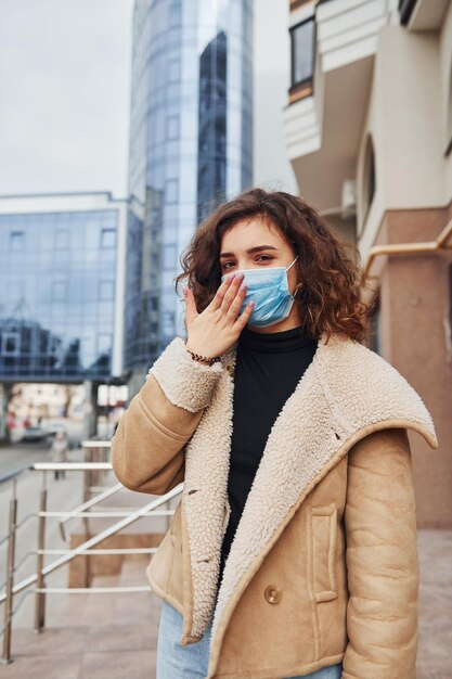 Portrait of young girl with curly hair in protective mask that have a cough outdoors near business building at quarantine time Conception of coronavirus